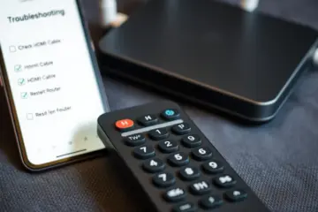 alt_text: A modern remote on dark fabric. A blurred phone shows a troubleshooting checklist. The mood is practical and solution-focused.