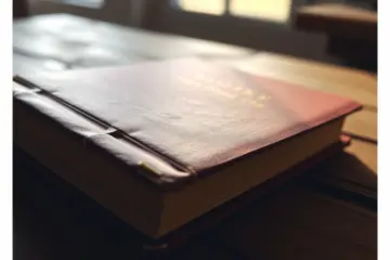 alt_text: A weathered burgundy book with a gold title rests on a rustic wooden table in soft morning light, evoking a quiet, timeless atmosphere.